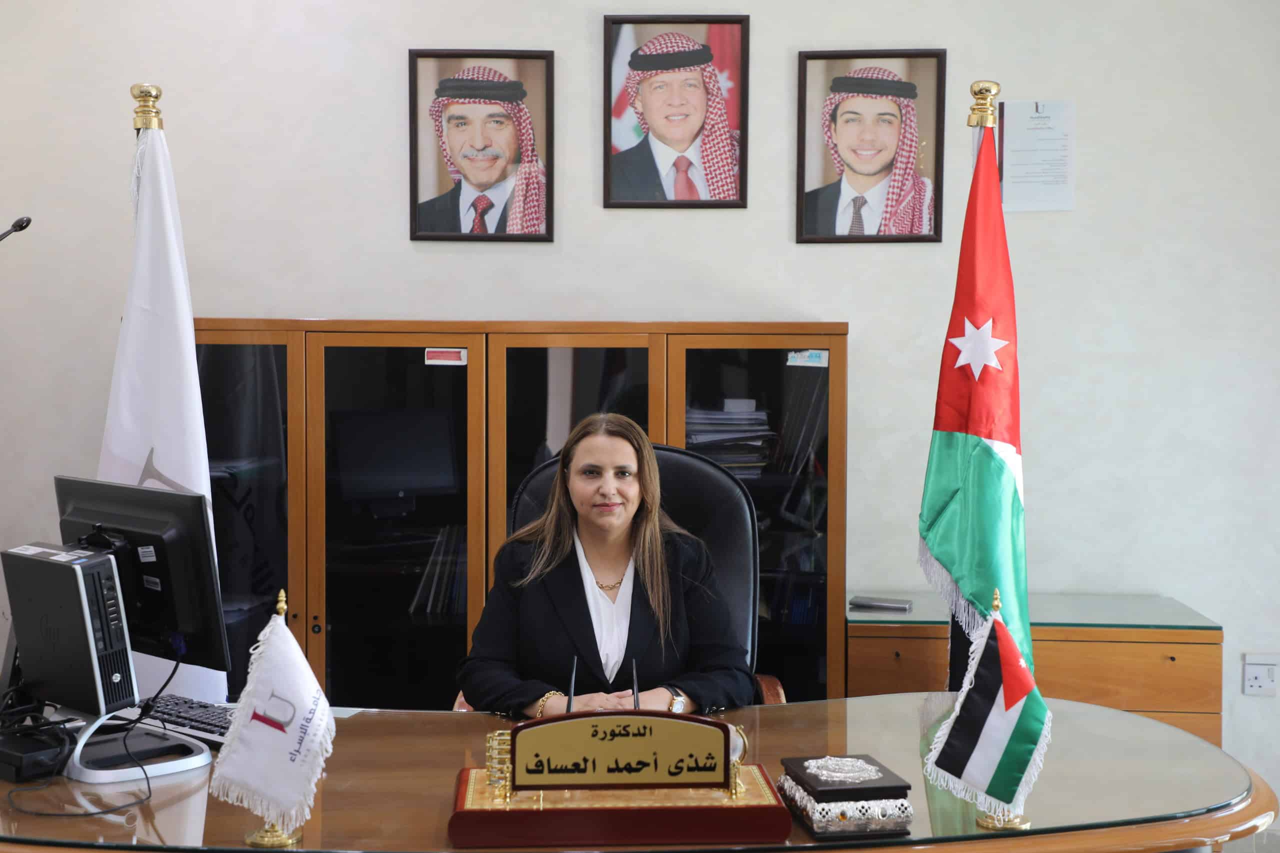 صورة لمديرة تجلس خلف مكتبها في جامعة الأردنية، مع صور لملوك الأردن على الحائط وعلم الأردن بجانبها.