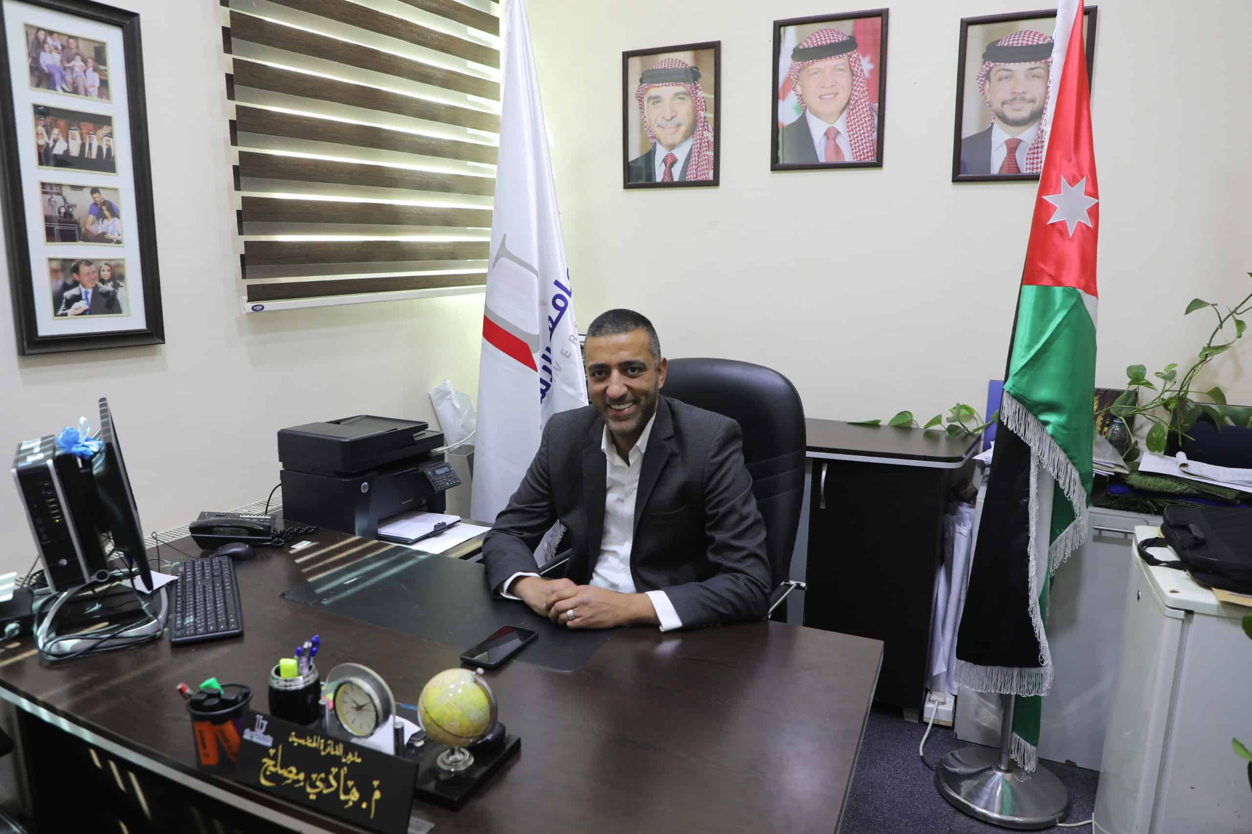 رئيس الدائرة الهندسية في جامعة الأردنية يجلس في مكتبه مع العلم الأردني والخلفية صور لطلاب وأعضاء هيئة التدريس.
