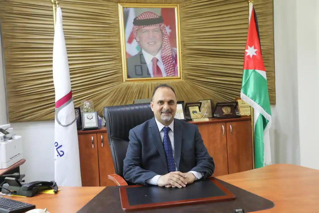 رئيس جامعة العلوم الإسلامية الأردنية يجلس في مكتبه مع صورة الملك عبد الله الثاني وراية الأردن في الخلفية.